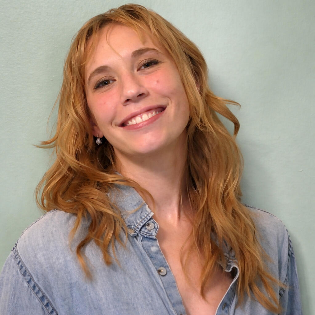 Artist Photo of Kestrel Preston Photo of the artist behind Sweetvine, Kestrel Preston, woman with strawberry blonde hair and blue blouse standing in front of a green wall